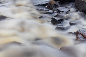 Close-up of a river flowing over rocks with a 13-second shutter speed, creating a mist-like effect. The water appears soft and ethereal, contrasting with the textured rocks. 