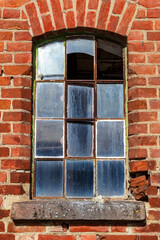 Old arched window in a red brick wall with a rusted iron frame, some glass panes missing or broken, revealing weathered decay and history