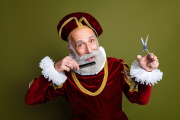 Elderly man dressed as a royal nobleman holding scissors and comb with a humorous pose against a...