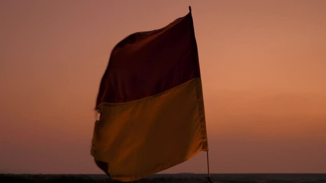 A yellow and red lifeguard flag flutters against a mesmerizing pink-orange sunset backdrop. The vivid colors of the flag and sky create a striking and serene coastal scene.