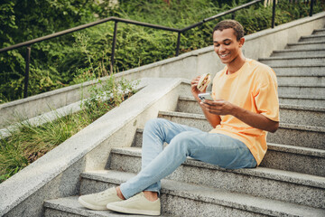 Obraz premium Young man with a trendy t-shirt enjoys a casual day outdoors while checking his phone and having a snack on urban steps in the city