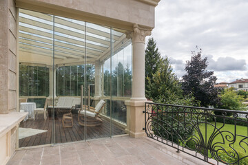 Fototapeta premium Glazed terrace of a luxurious country mansion decorated in neoclassical style. Stylish furniture. Beautiful view of the green lawn with trees.