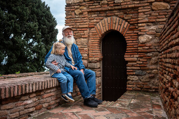 Fototapeta premium Grandfather and granddaughter sitting together on a brick wall, enjoying a peaceful moment with a view of a rustic brick archway and door in the background