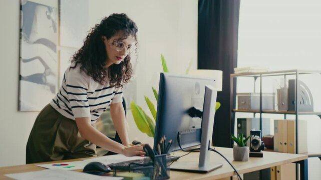 Cute Caucasian female wearing glasses for better vision while leaning over office desk and writing with pencil. Taking notes or organizing task while working in bright modern workspace.