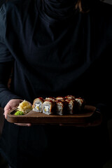 A skilled hand holds a wooden platter adorned with a vibrant array of sushi rolls topped with savory sauces, complemented by fresh wasabi and pickled ginger, creating an inviting dining experience