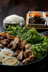 A delicious platter featuring grilled meat, fresh lettuce, and onions is artfully arranged on a black sizzling plate. Accompanying dishes of rice and pickled sides create a harmonious meal