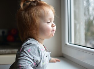 Red messy hair toddler girl curiously looking through the window. Daughter impatiently waiting...
