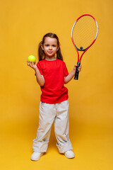 Happy little sports girl in red t-shirt holding tennis racket and tennis ball on yellow background....