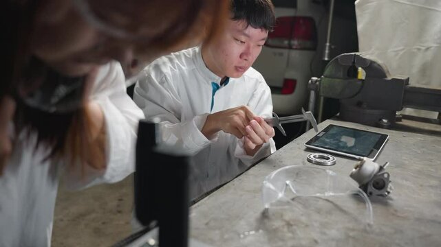 Partial view of lady putting on face shield while observing microscope and young technician adjusts vernier caliper, taps on tablet in lab with carburetor on table and white car parked in background