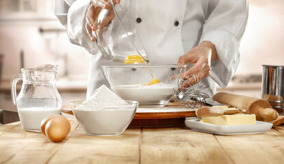 Person in a chef's uniform at wooden kitchen countertop pouring an egg into large glass mixing bowl...