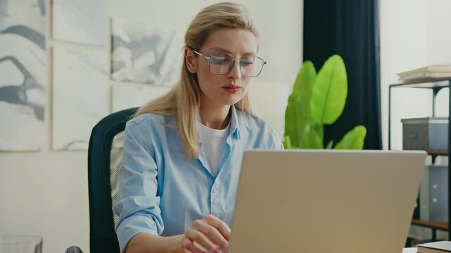 Thoughtful Caucasian woman with blond hair sitting at desk while resting chin on hand. Looking to side while analyzing information or considering ideas. Appearing focused and engaged.
