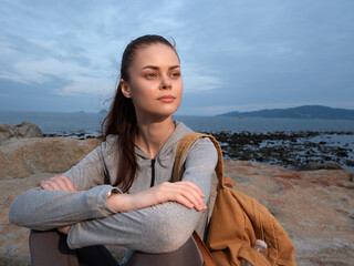 Young woman with a backpack sitting on rocks by the sea, looking thoughtfully at the horizon during...