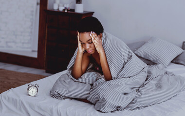 Insomnia concept. African american girl in blanket sits on bed with alarm clock, dark