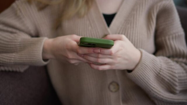 Close-up of young lady operating iPhone with green case while seated at wooden table, her hands are focused on screen, showing attention to detail in a relaxed environment