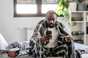 Sick man with a plaid blanket, wearing pajamas, sitting on a bed, checking his phone while holding...