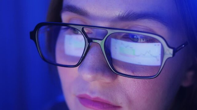 Trader woman wearing glasses working on laptop close-up footage. Screen with stock market charts and reflection of Bitcoin or data analysis. High quality 4k footage