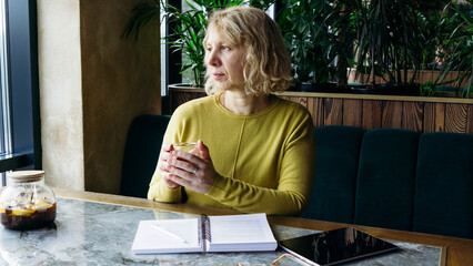 A senior businesswoman enjoys a beverage while using a tablet, embracing the freelance remote work concept. Her relaxed demeanor in a modern workspace reflects the flexibility and freedom of digital e