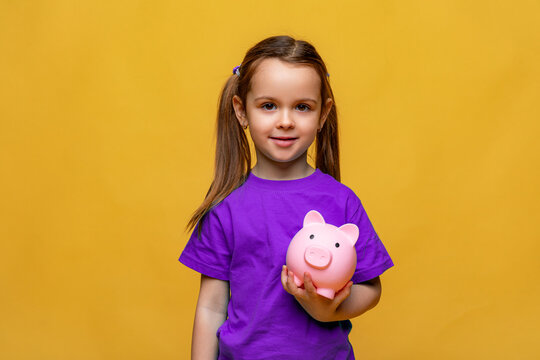 Happy little girl in purple t-shirt holding pink piggy bank money box on yellow background. Financial concept of children pocket money. Save money for education and family expenses