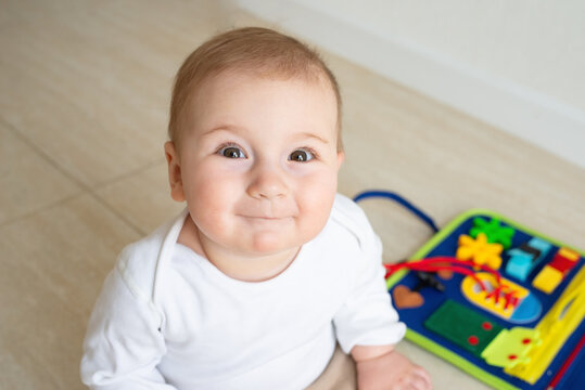 Cute baby girl smile sitting on the floor. On the background developing bright handmade toy for fine motor skills made of felt. Childhood development, soft busy book with laces, zippers and Velcro.