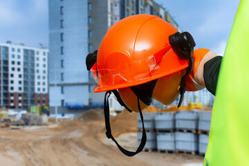 Construction worker in a high-visibility vest holding an orange safety helmet and protective gear at a building site.