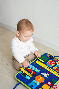 Baby girl sit plays on the floor with a developing bright handmade toy for fine motor skills made of felt. Early childhood development, soft busy book with with laces, zippers and Velcro