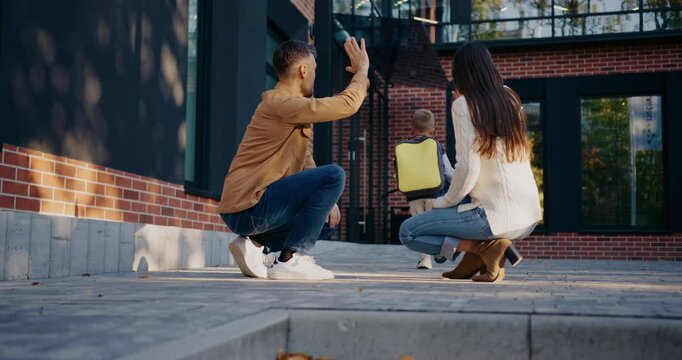 Small Caucasian boy giving high five to his dad and mom. Running away towards school entrance. Carrying square yellow backpack. Parents sitting down and waving with their hands to beloved child.