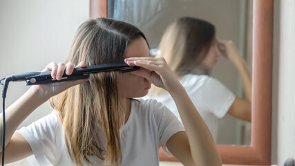 Fototapeta premium Young woman using a hair iron and doing a hairstyle in the morning at bedroom at home
