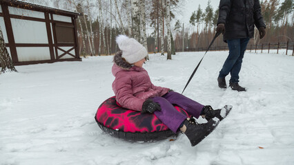 Father pulling child girl sitting on inflatable tube in snow (inflatable sled)