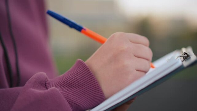 Close-up side view of individual in maroon top taking notes on clipboard outdoors, using an orange pen, background shows sports field with people walking
