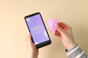 Menstruation. Woman holding smartphone with period calendar application and menstrual cup on beige background, top view
