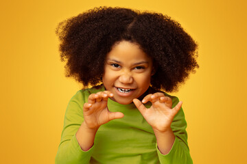 Playful little black girl imitating tiger, roaring and grimacing over yellow studio background