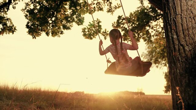 Funny little girl plays swings under old tree at sunset time. Happy little girl enjoys flight feeling on swings in country field on summer holiday. Little girl rides handmade swing in country garden
