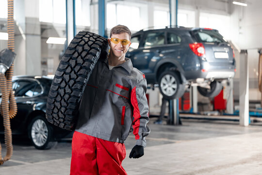 Happy male auto mechanic carrying tire in garage workshop, seasonal wheel change