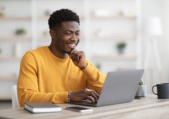 Happy black guy project manager sitting at workdesk at office, typing on laptop keyboard and...