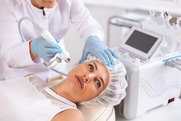 Young adult woman undergoing skin therapy session in modern cosmetology clinic. Aesthetician applying atomized nutrients with airbrush onto client face to enhance and revitalize complexion