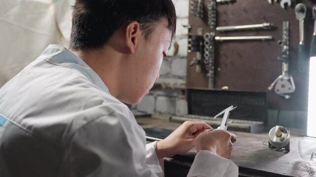 Young engineer working with veiner caliper, adjusting and measuring diameter of boris in hand, in front of bright light, background features mechanical tools, workbench, and precision instruments