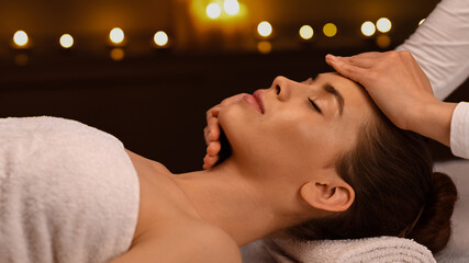 Sleeping young woman getting healing face massage at modern luxury spa in romantic atmosphere, side...