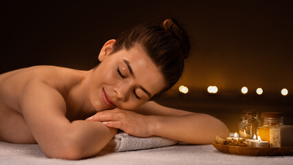 Side view of tranquil young woman spending day at spa salon decorated with candles, getting aroma session with relaxing full body massage, panorama with copy space. Spa, massage, relaxation concept