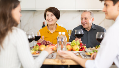 Positive mature couple celebrating family holiday with son and his young wife at kitchen table with wine and light snacks