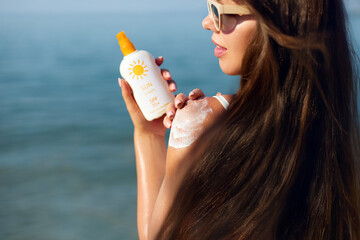 A woman enjoys a sunny day at the beach, applying sunscreen to her shoulder. Girl wears sunglasses...
