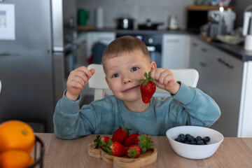 Happy little boy sitting at wooden table in kitchen, holding fresh strawberry and smiling joyfully. Concept healthy eating, fresh organic food, and balanced nutrition. High quality photo