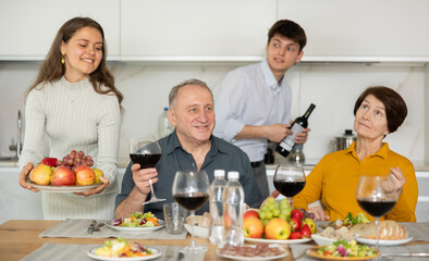 Happy carefree senior parents sharing memories with grown daughter and son-in-law during conversation over family celebration with wine at cozy home environment