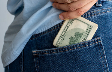 Close-up of person wearing blue jeans and light blue shirt putting stack of US dollar bills into...