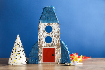 Beautiful pinata in shape of rocket, party cones and blowers on wooden table against blue background