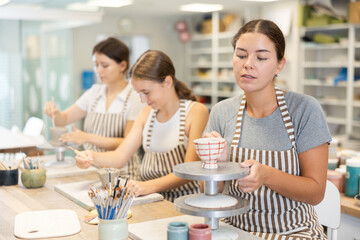 In pottery workshop, three girls are sitting at table, performing sculpting and coloring products made of raw clay. Amateurs are busy with hobbies leisure activity at master class