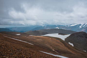 Dark overcast landscape with high pass, wide glacier on stony hill slope and snowy mountain range silhouette in far away in rainy low clouds. Large mountains with snow under gray sky in bad weather.