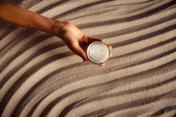 Hand holding a jar of skincare cream on a textured striped surface emphasizing natural beauty and...