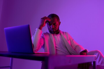 Thoughtful man in a stylish suit staring at a laptop in colorful lighting