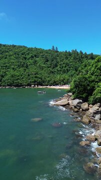 Aerial view of Praia do Cedro located in Ubatuba, Brazil. People enjoy the beach and the sea water crashes against the rocks on the coast. Green trees are seen in the beautiful landscape.
