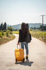 Musicien anonyme de dos, marchant sur une route de campagne avec son étui de guitare dans le dos et une valise à roulette jaune